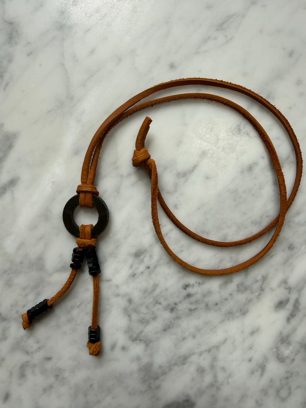 The black necklace on marble from a second angle, showing the knot that closes the leather strap and the black nuts slid apart along both strands.