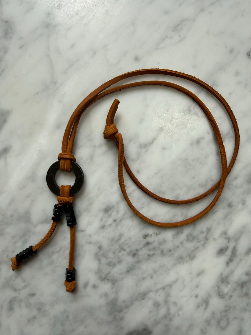 The black necklace on white marble. A brown leather strap is coiled above; a black steel washer sits at the centre with black hex nuts spaced along the two strands below it.