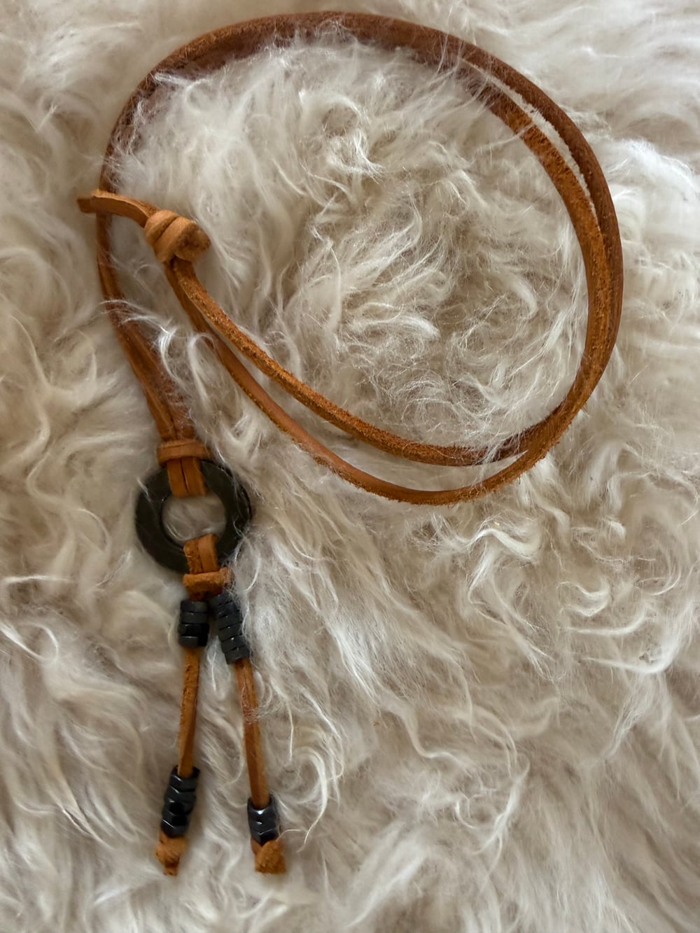 The black necklace resting on cream sheepskin, the black washer and black hex nuts against the brown leather strap.