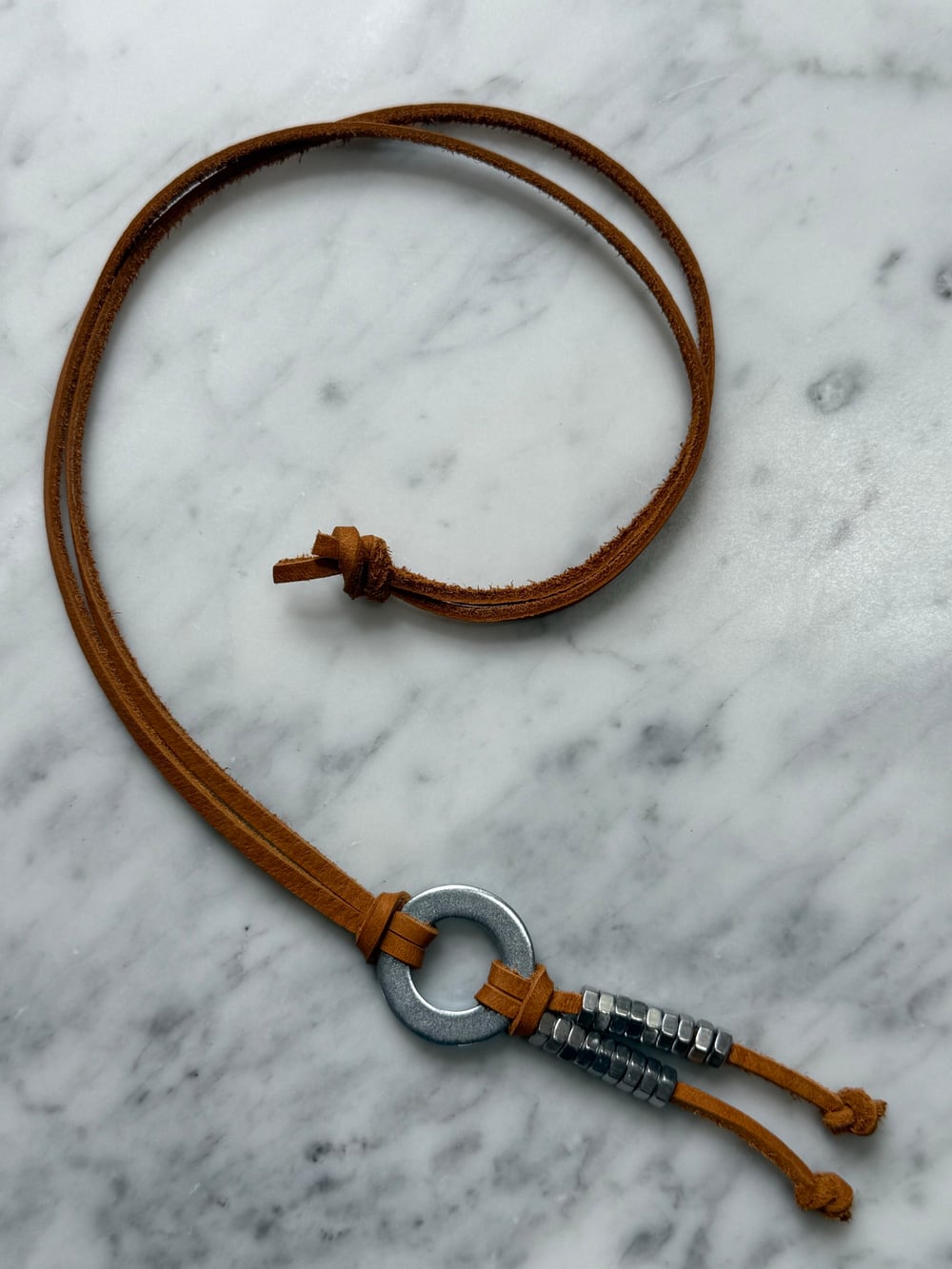 The silver necklace on marble from a second angle, with the leather strap looped and the washer and hex nuts in the lower right.