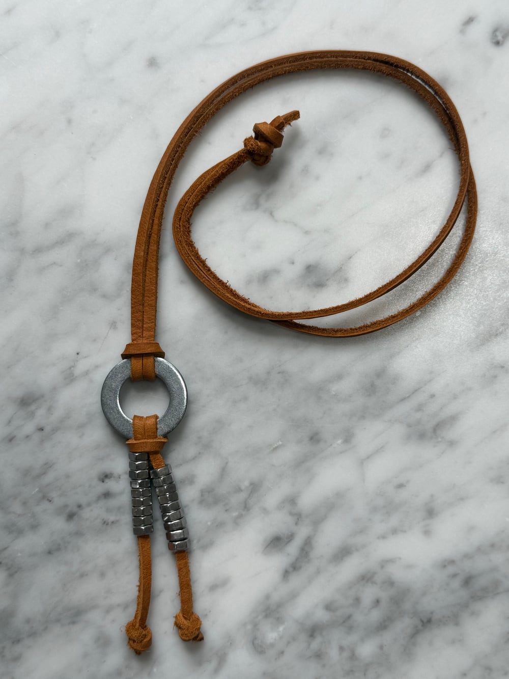 The silver necklace laid out on white marble. A brown leather strap is coiled to the left; a round steel washer sits at the centre with two short strands of small hex nuts hanging from it.